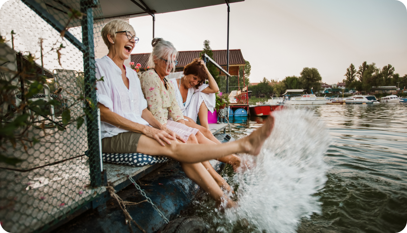 Women’s Health Blog In Text 3 A group of older women sitting on a dock, smiling, and splashing their feet in the water.